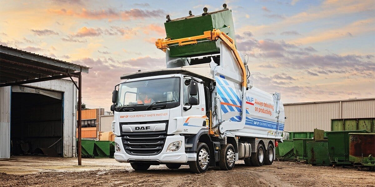 European DAF cab-over front loader garbage truck lifting dumpster at industrial site, showing modern waste collection design and driver-first cab layout.