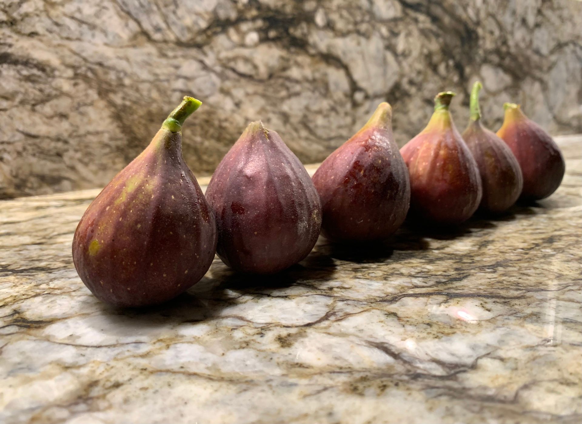 A half-dozen dark plum-colored figs sit in a row upright on a granite counter.