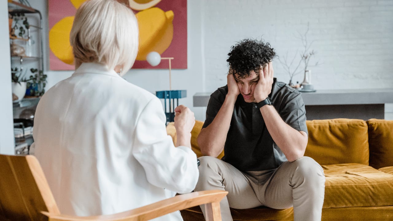 Man sitting on couch with hands on face, while a person sits across from him.
