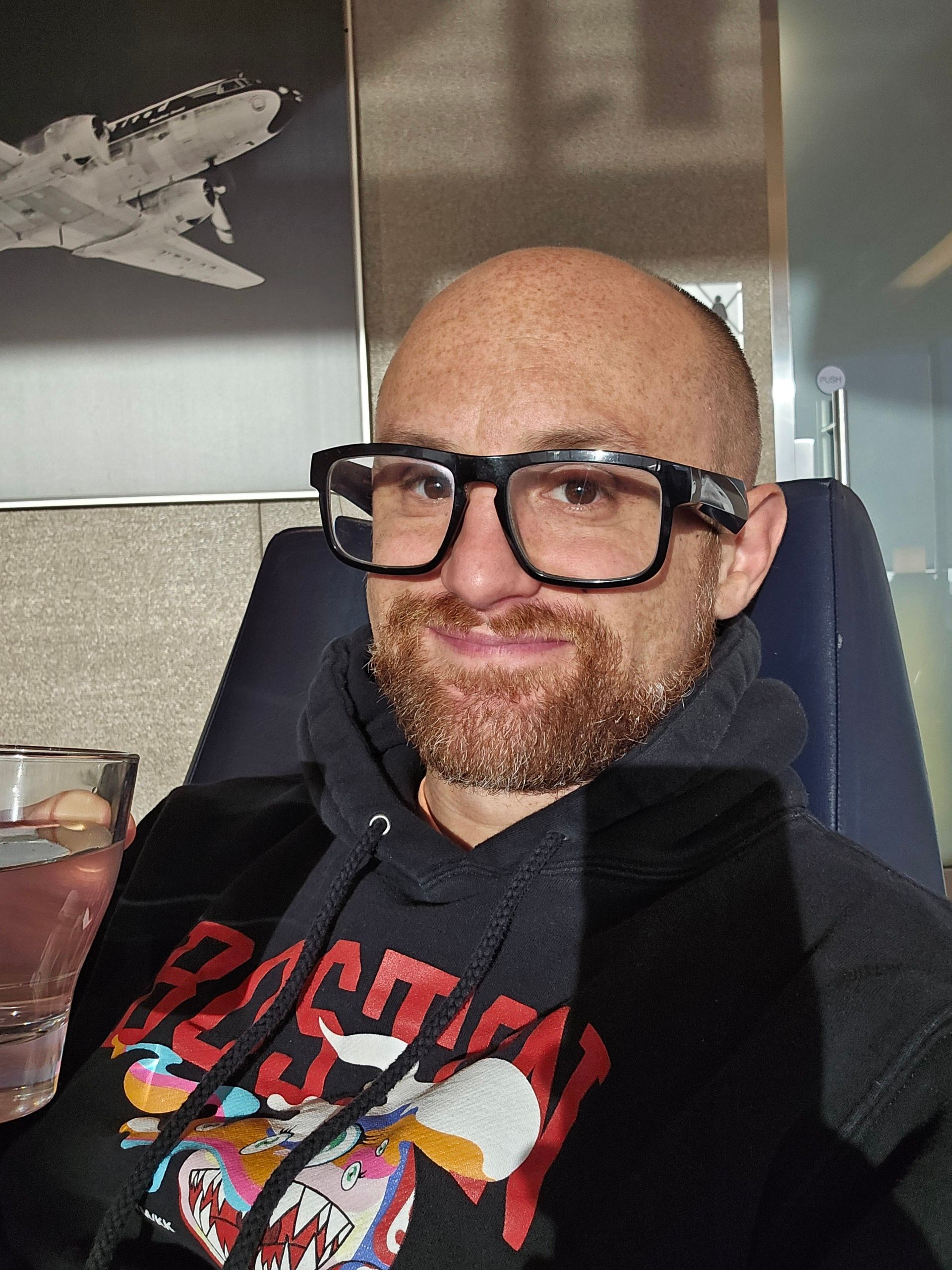 A selfie of me in a seat in an airport lounge, holding a glass of water, with a black and white photo of a United plane flying against a grey sky in the background. I'm a bald, middle-aged, white man with a red beard flecked with white. I'm wearing glasses with thick black rims and a black sweatshirt with Boston written in red across the chest with a Murakami monster below.