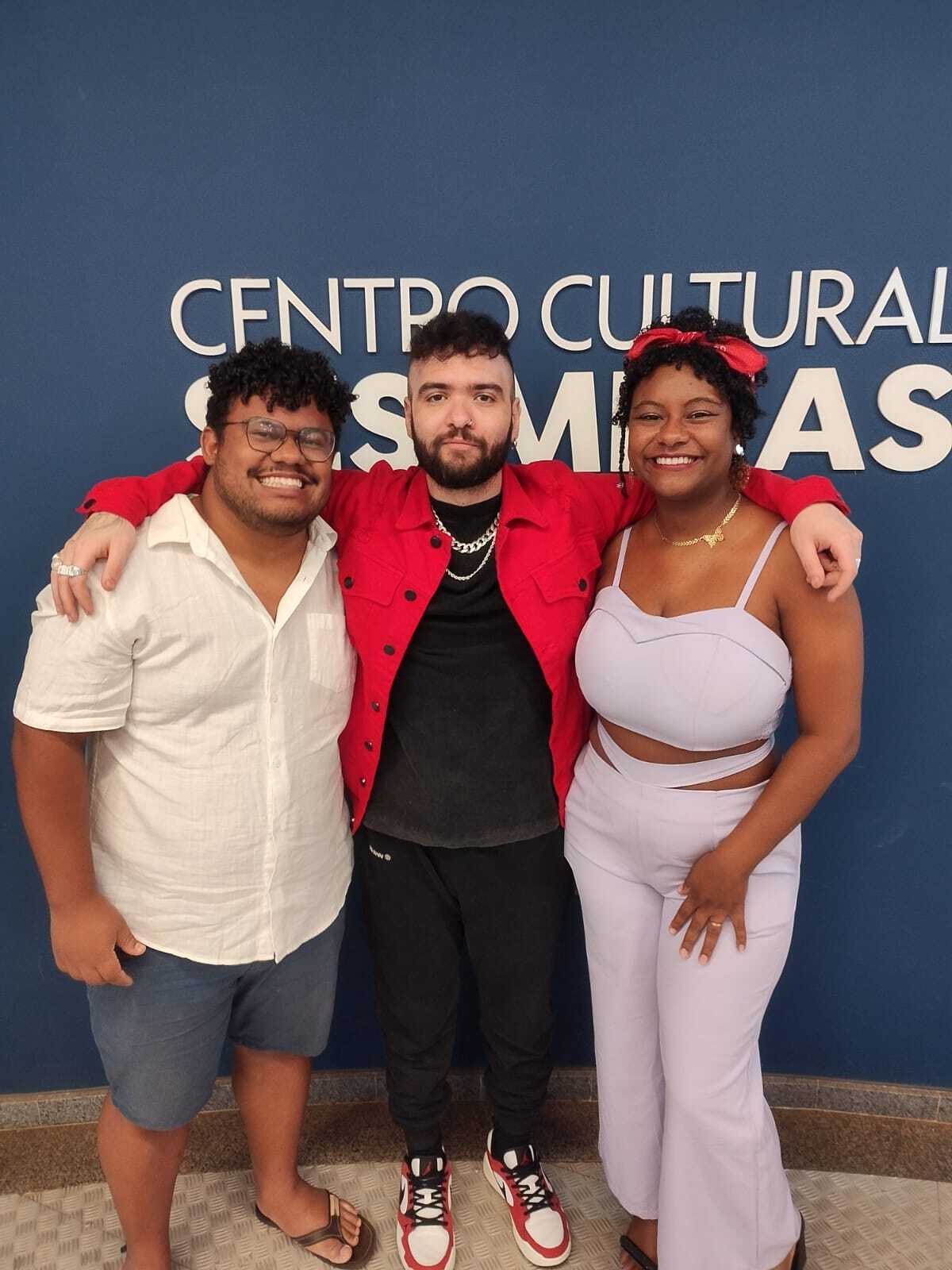Duas pessoas negras, felizes, sorrindo e abraçando o mestre Tiago Santinelli. Da esquerda para a direita; Ícaro, homem negro, de óculos e cabelos cacheados, vestindo uma camiseta de botões branca e short azul. Ao seu lado, Tiago Santinelli, comediante e militante, homem branco com barba, camisa e calça preta. Por fim, Vanessa, esposa de Ícaro, mulher negra com cabelo crespo preso a uma bandana vestindo um cropped e calça lilás.