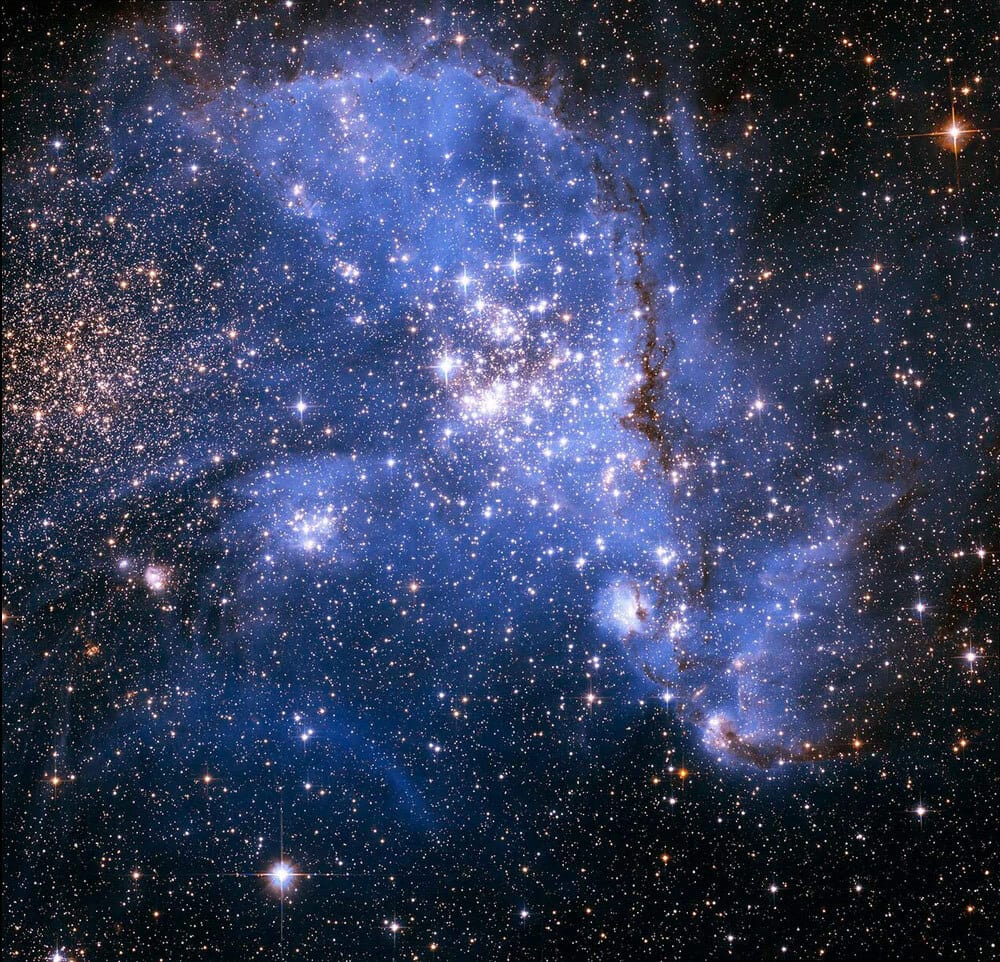A spectacular cluster of brilliant stars sits in a cloud of bluish gas.