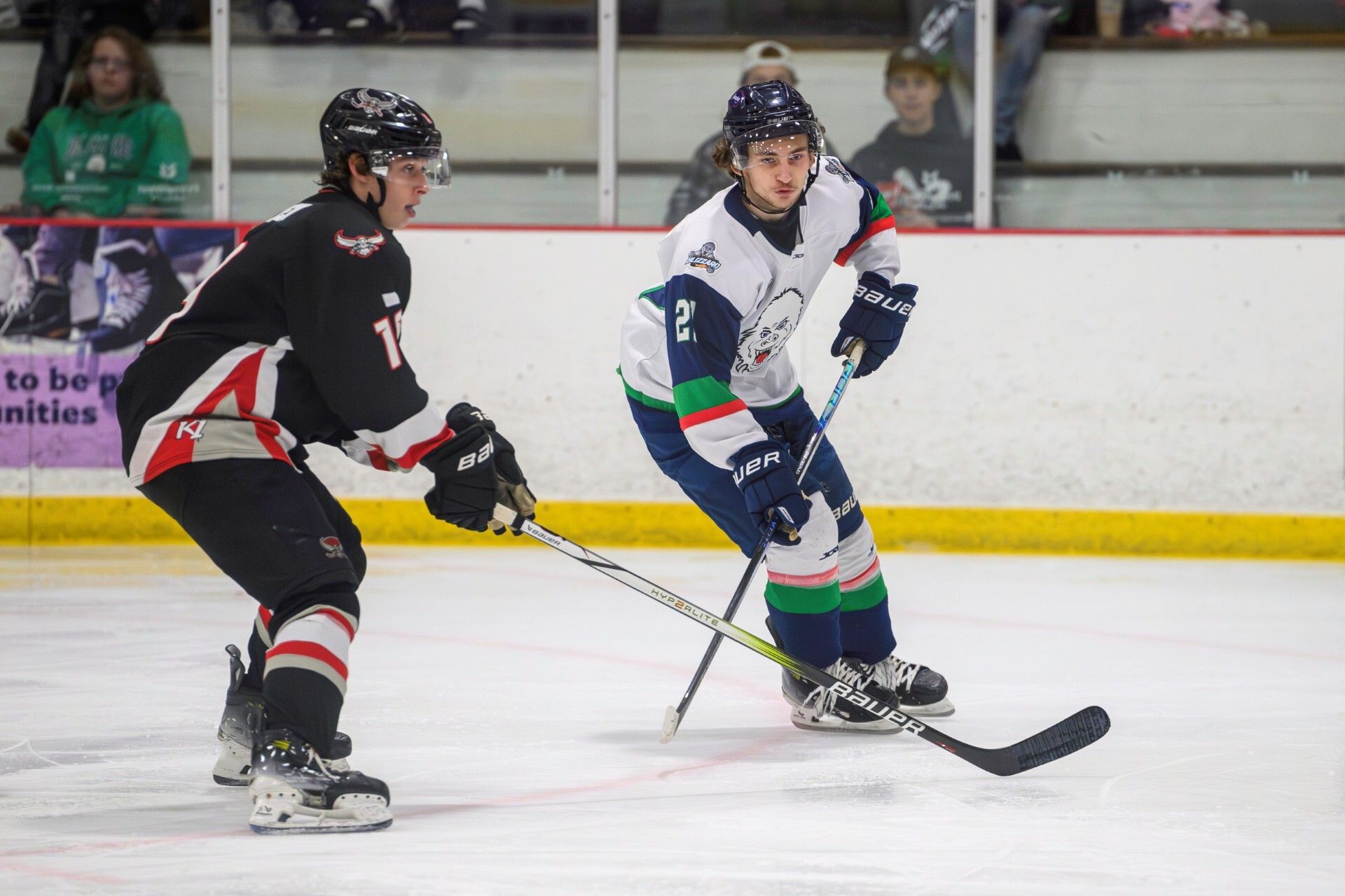 A men's hockey player in a dark uniform and a men's hockey player in a white uniform skate during a recent game. 