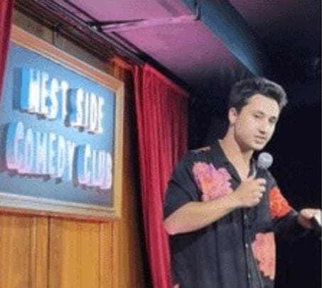 A man in a floral shirt performs on stage with a microphone at the West Side Comedy Club, with a neon sign and red curtains in the background.