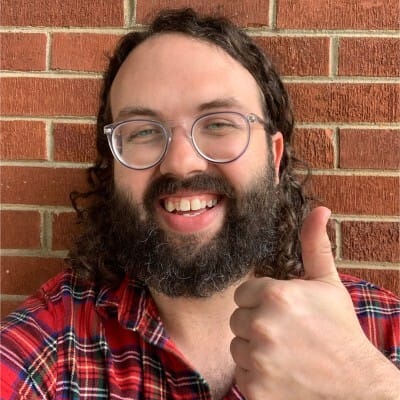 A smiling Travis Hazelwood, with glasses, long curly hair, and a beard giving a thumbs-up gesture while standing in front of a red brick wall.