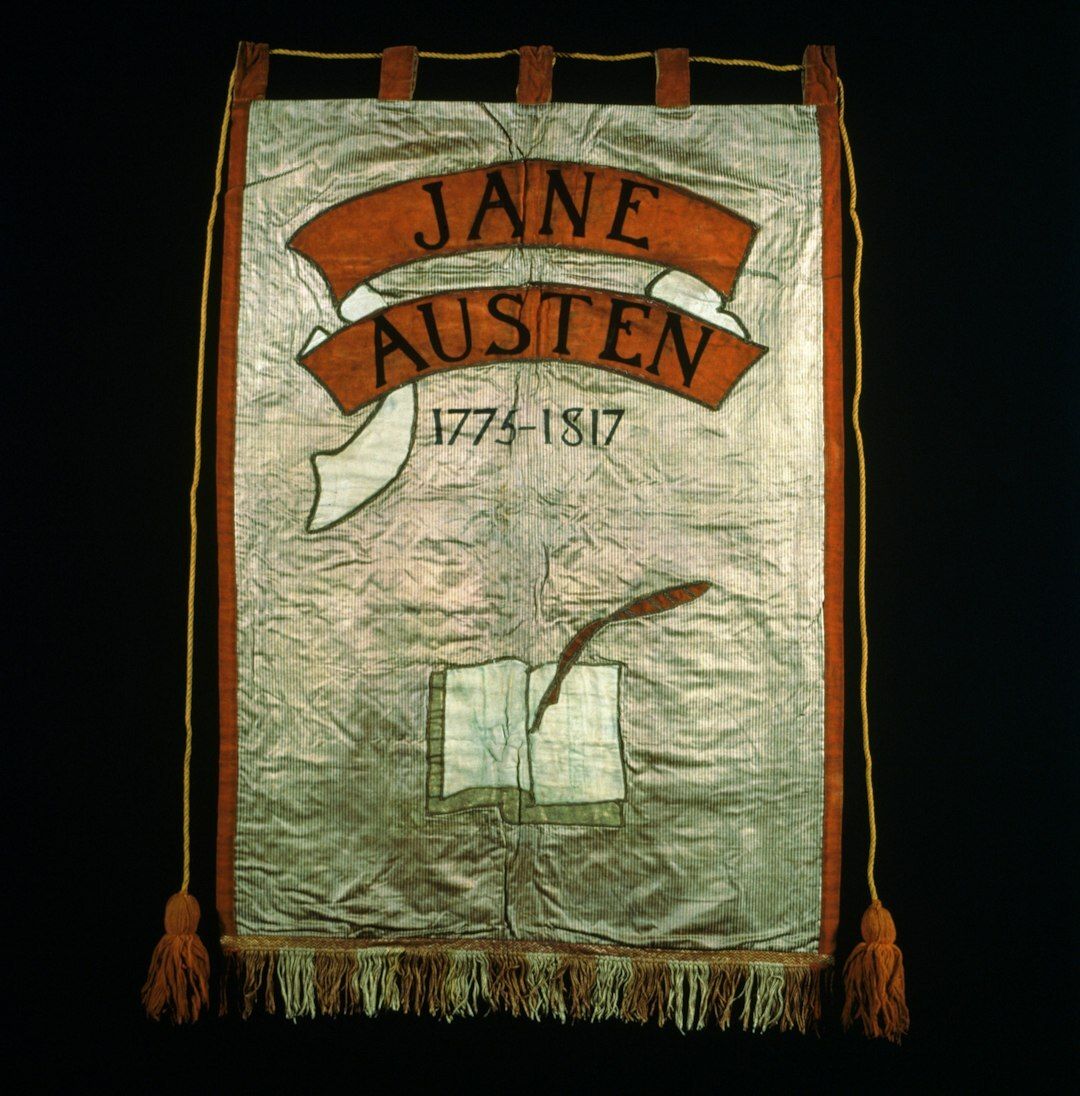 Jane Austen Suffrage Banner, 1908. Artist: Mary Lowndes. Cream yellow and red silk satin ground with black painted lettering and orange and cream appliqued scrolls and orange painted lettering. Cream wool book design. Yellow silk wrapped cords and orange tassels. F06DMAST