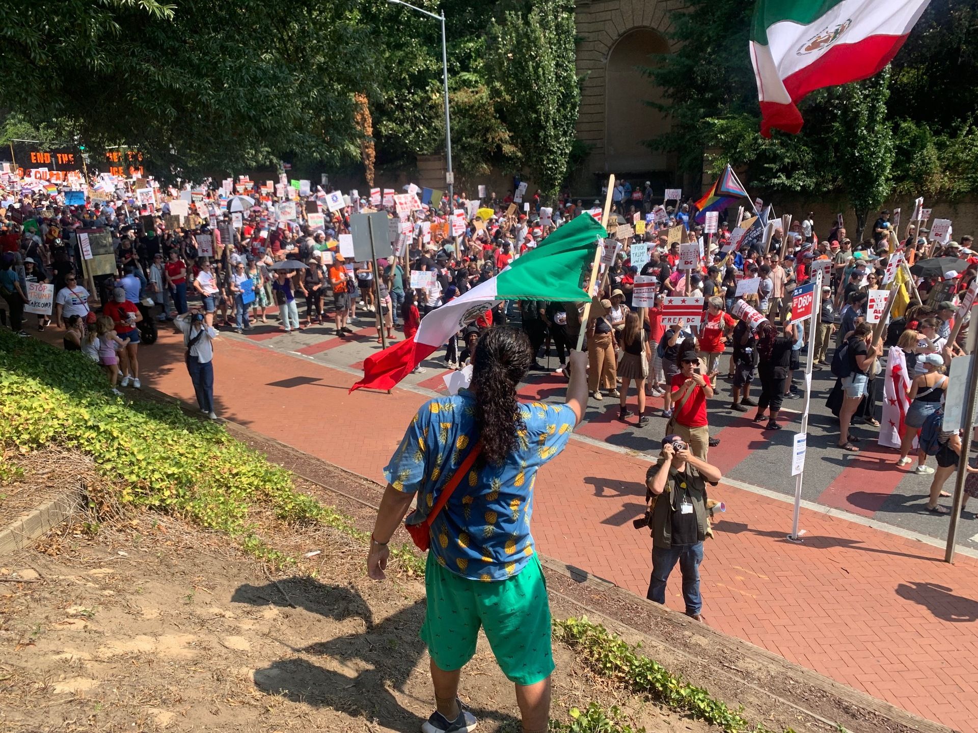 Someone with long dark hair in a ponytail is seen from behind, as he waves a Mexican flag on the slope above 16th Street NW. A crowd of demonstrators fills the street. Below, a photographer is taking a picture.