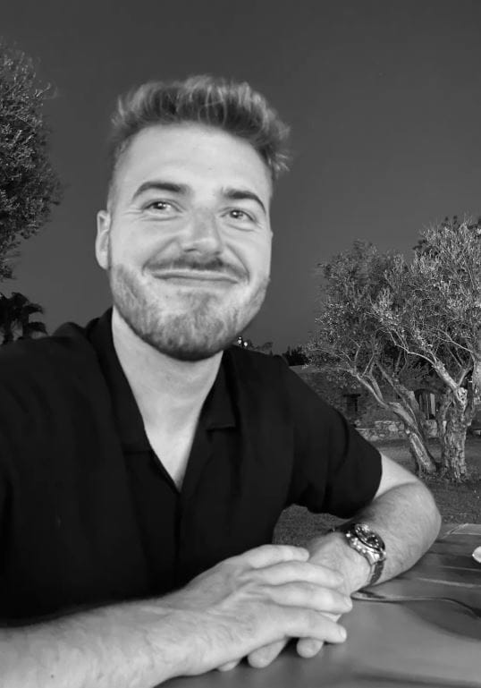 Black and white photo of a young man looking straight into the camera. He has a beard and he is smiling. He is wearing a black t-shirt and a watch. Trees in the background.