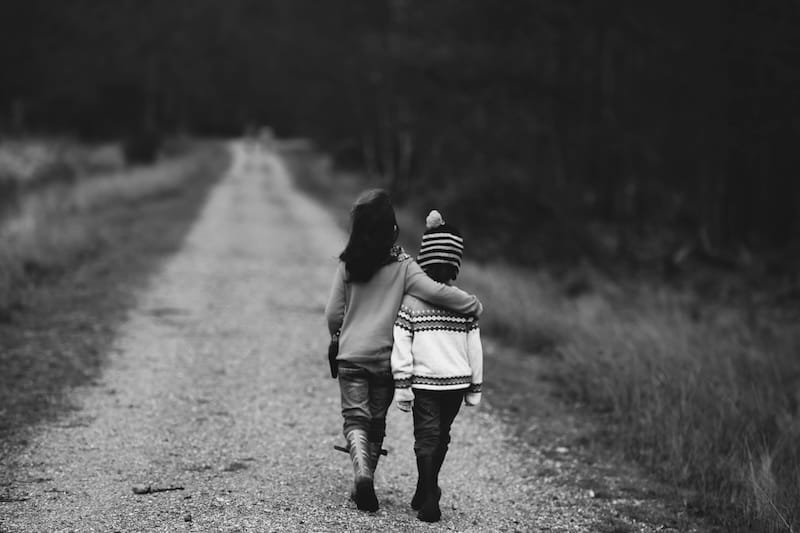 This black-and-white photograph shows two children walking away from the camera down a gravel or dirt road. They are both wearing light-colored sweaters. The child on the left is slightly taller and has long dark hair. THe child on the right is wearing a woven hat. The child on the left has their arm around the child on the right.