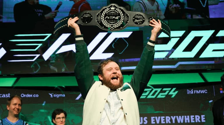 A bearded man on stage holding a wrestling-style championship belt above his head, smiling as a crowd and green stage lights shine behind him, celebrating his victory at a competitive spreadsheet event.
