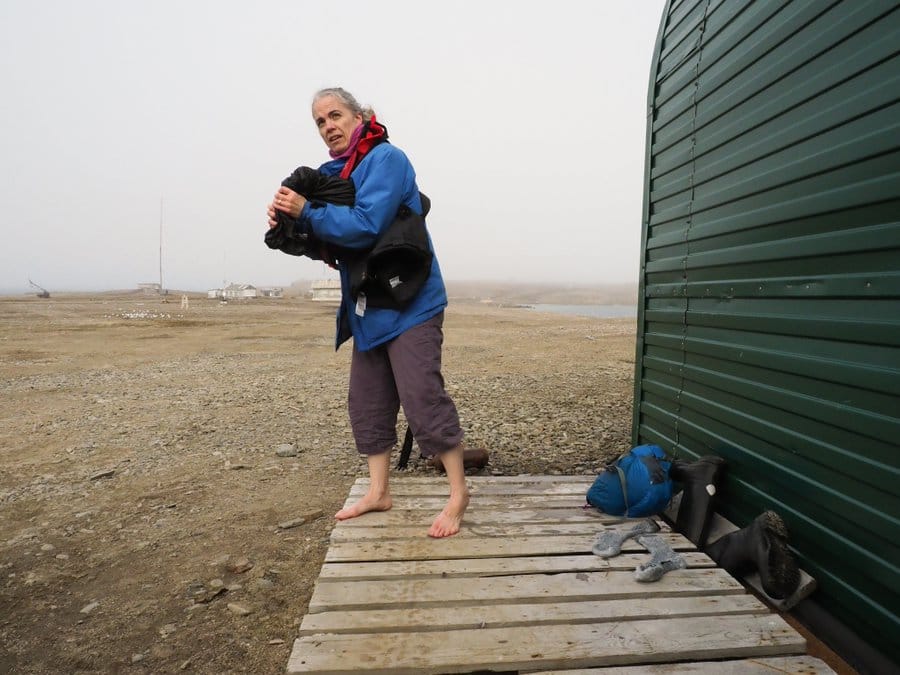 Photo of a woman with gray hair standing barefoot in a flat and rocky landscape wringing out something black. A prefabricated wall of a metal hut is visible at right. Wet socks are lying next to the woman on a wooden pallet, and a pair of boots are turned upside down. 