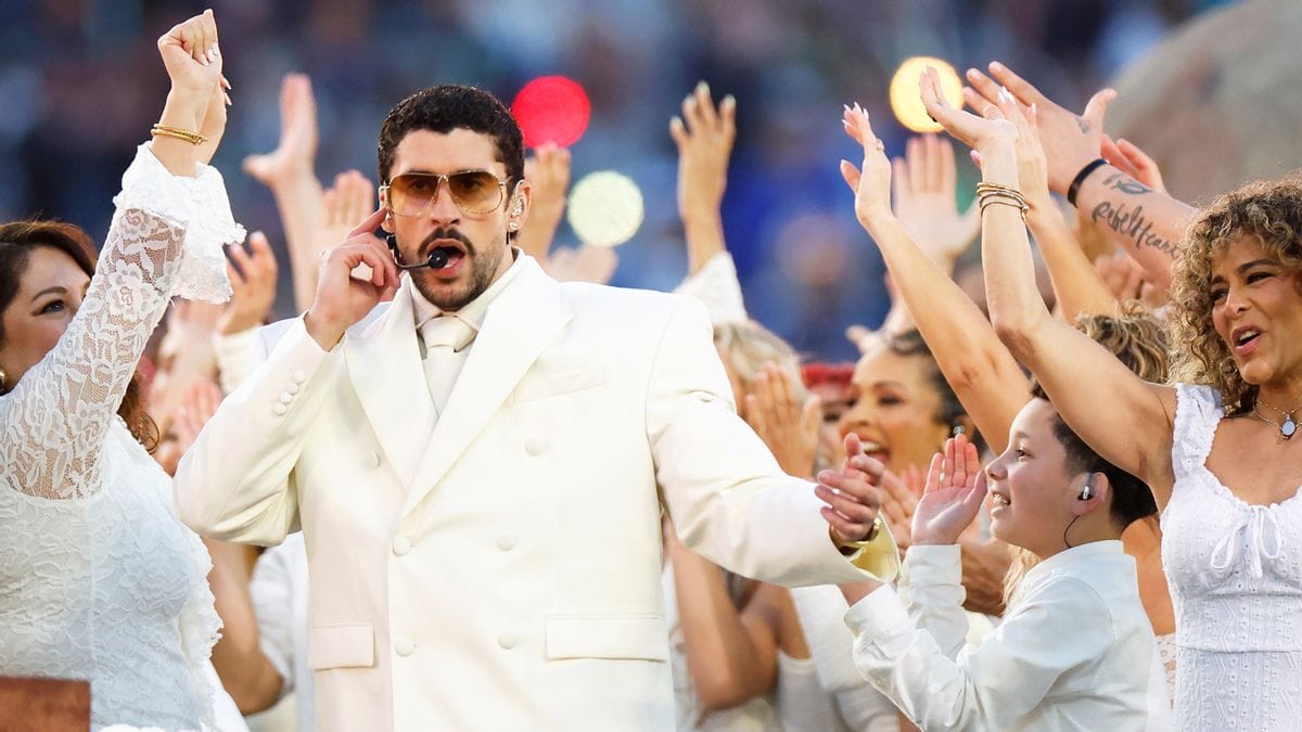 A Latino wearing a white suit sings surrounded by other people wearing white outfits who are smiling.