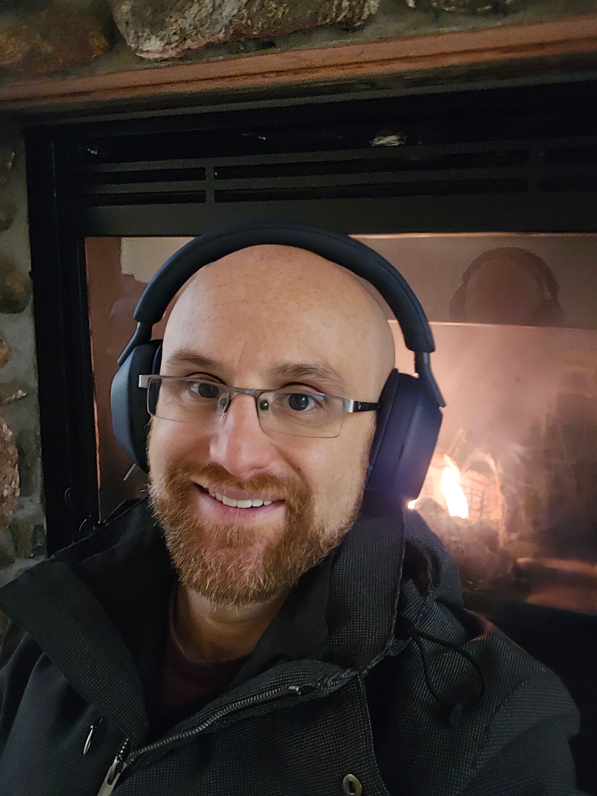 A selfie of me in front of a glass-covered fireplace in a stone chimney with a fire burning amidst logs. I'm a bald, middle-aged, white man with a red beard flecked with white. I'm wearing dark blue over-ear headphones, glasses with metal top rims, and a dark blue jacket with a check pattern.