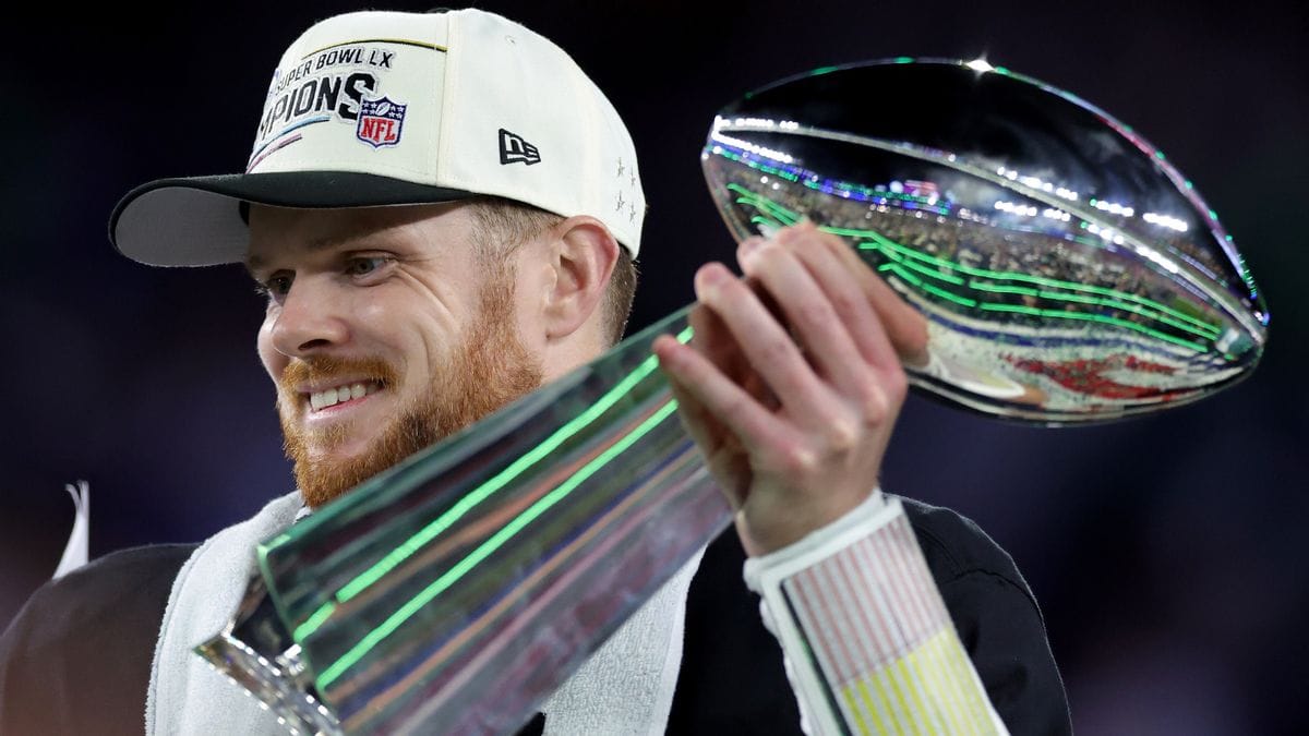 A white man wearing a white SUPER BOWL LX CHAMPIONS hat holds a large silver award.
