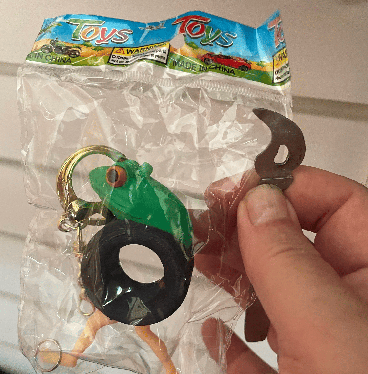 Bruce Sterling’s hand holding a packaged 3d-printed AI slopject that appears to be a frog head on the body of a car tire, with human legs. It’s also a keychain, I guess?