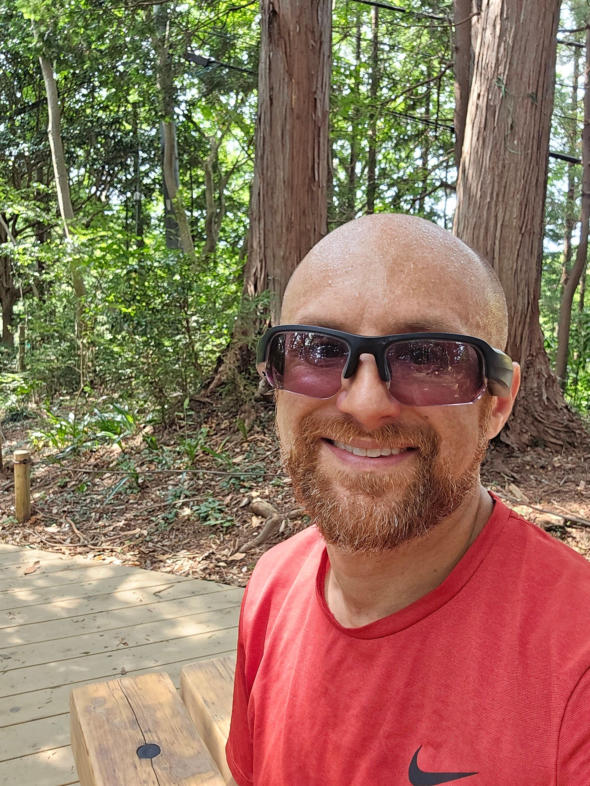 A selfie of me in a forest, with two large Japanese cypress trees behind me on a bright day. I’m sitting on a wooden bench on a wooden platform. I’m a bald, middle-aged, white man with a red beard flecked with white. I’m wearing black sunglasses and a red Nike running shirt.
