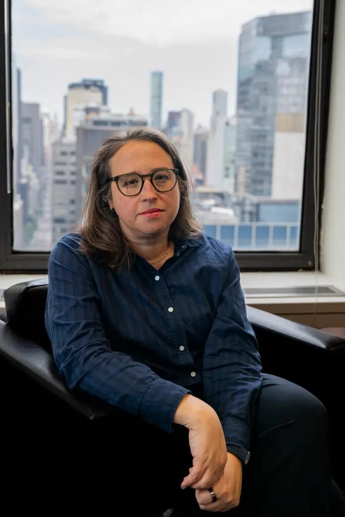 A person with long brown hair, glasses, and a dark blue button-down shirt sits in a black chair near a window overlooking tall city buildings.