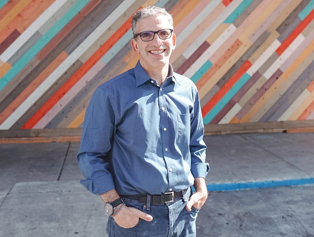 Alastair Goldfisher, contributing writer at AiNews.com, standing in front of a colorful diagonal wood-paneled wall, wearing a blue button-down shirt and glasses.