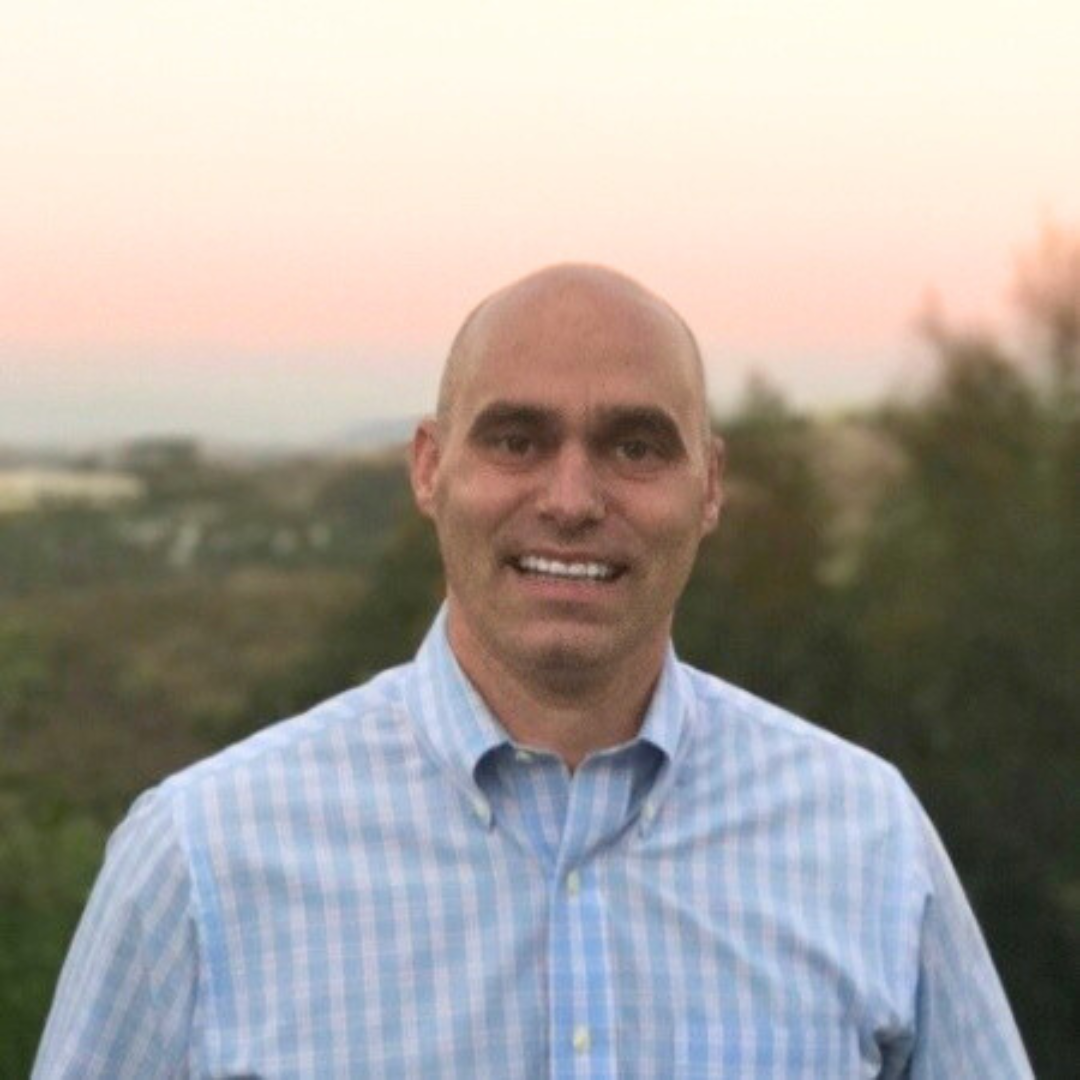 Michael Mazzotta, Founder of AiNews.com, smiling outdoors in a checkered shirt with a sunset landscape in the background.