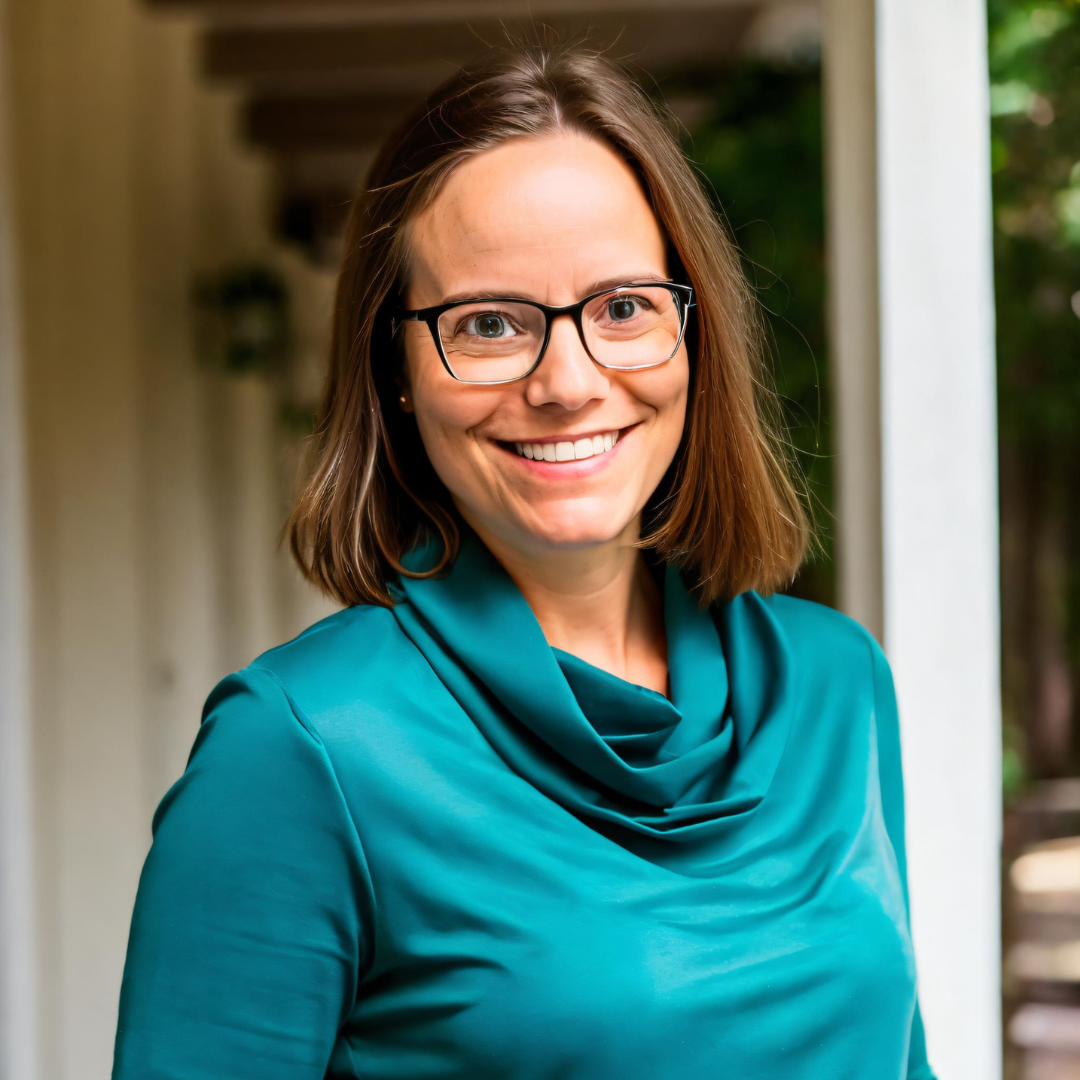Alicia Shapiro, Co-Founder and CMO of AiNews.com, smiling in a teal top and glasses, standing under a shaded outdoor walkway.