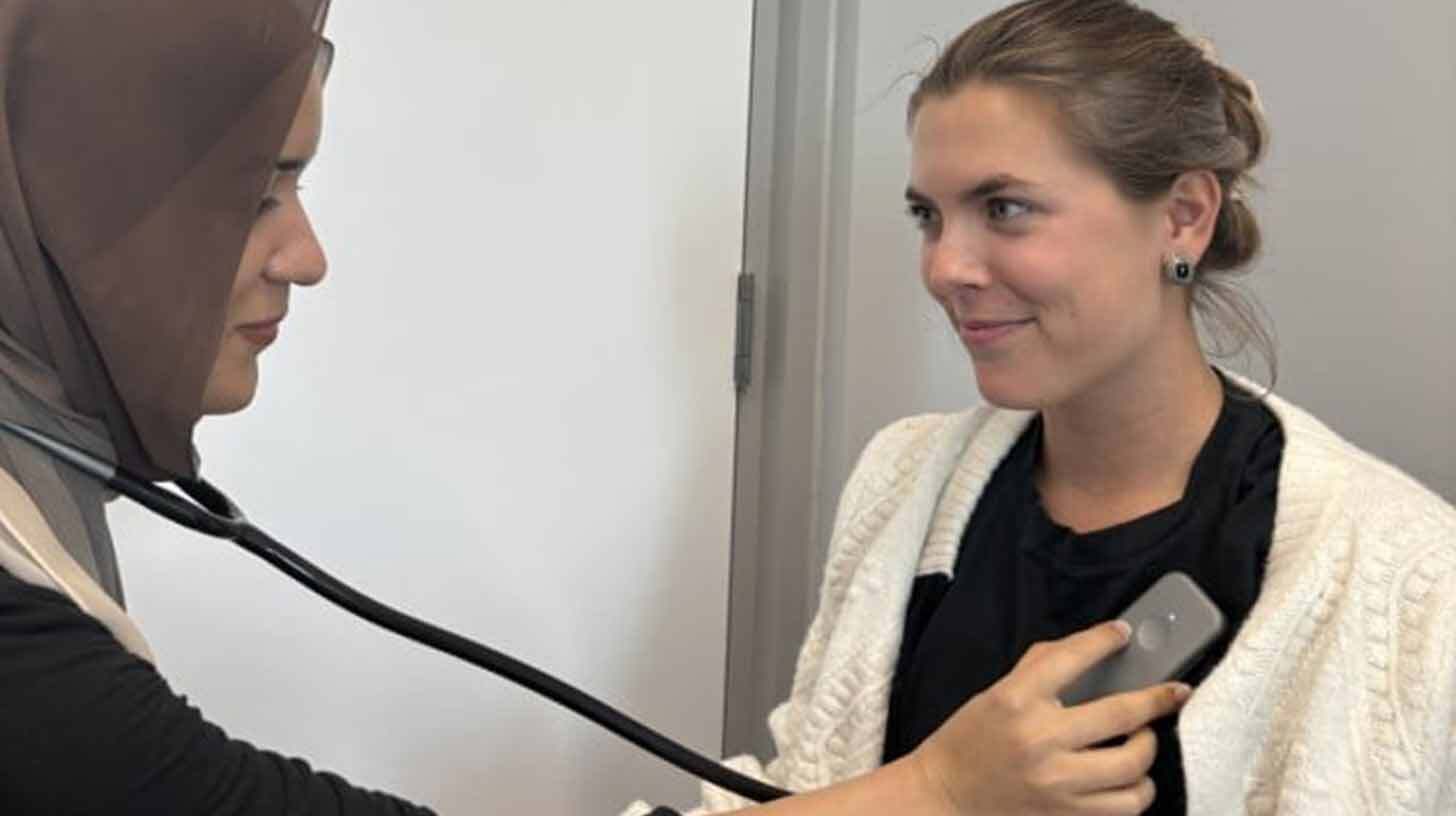 Kaks naist, üks kannab pead loorituna ja kasutab stetoskoopi teise naise südame kuulamiseks. Teine naine naeratab ja kannab valget kampsunit.
