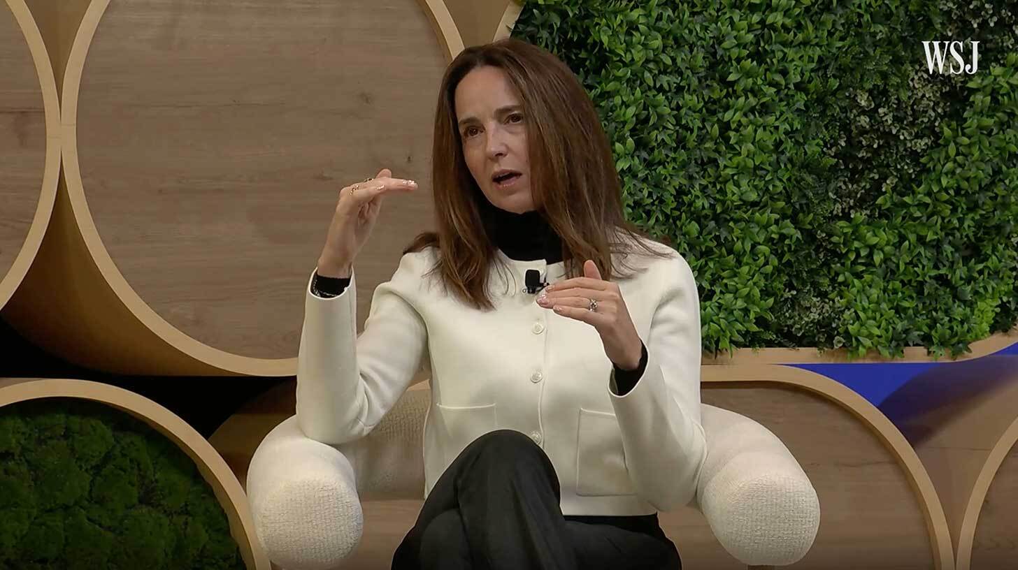 Naine istub toolil ja räägib käega žestikuleerides. Ta kannab valget jakki ja musta pükse. Taustal on suur puidust dekoratsioon ja rohelised taimed. Ülemises paremas nurgas on väikeses kirjas 'WSJ'.