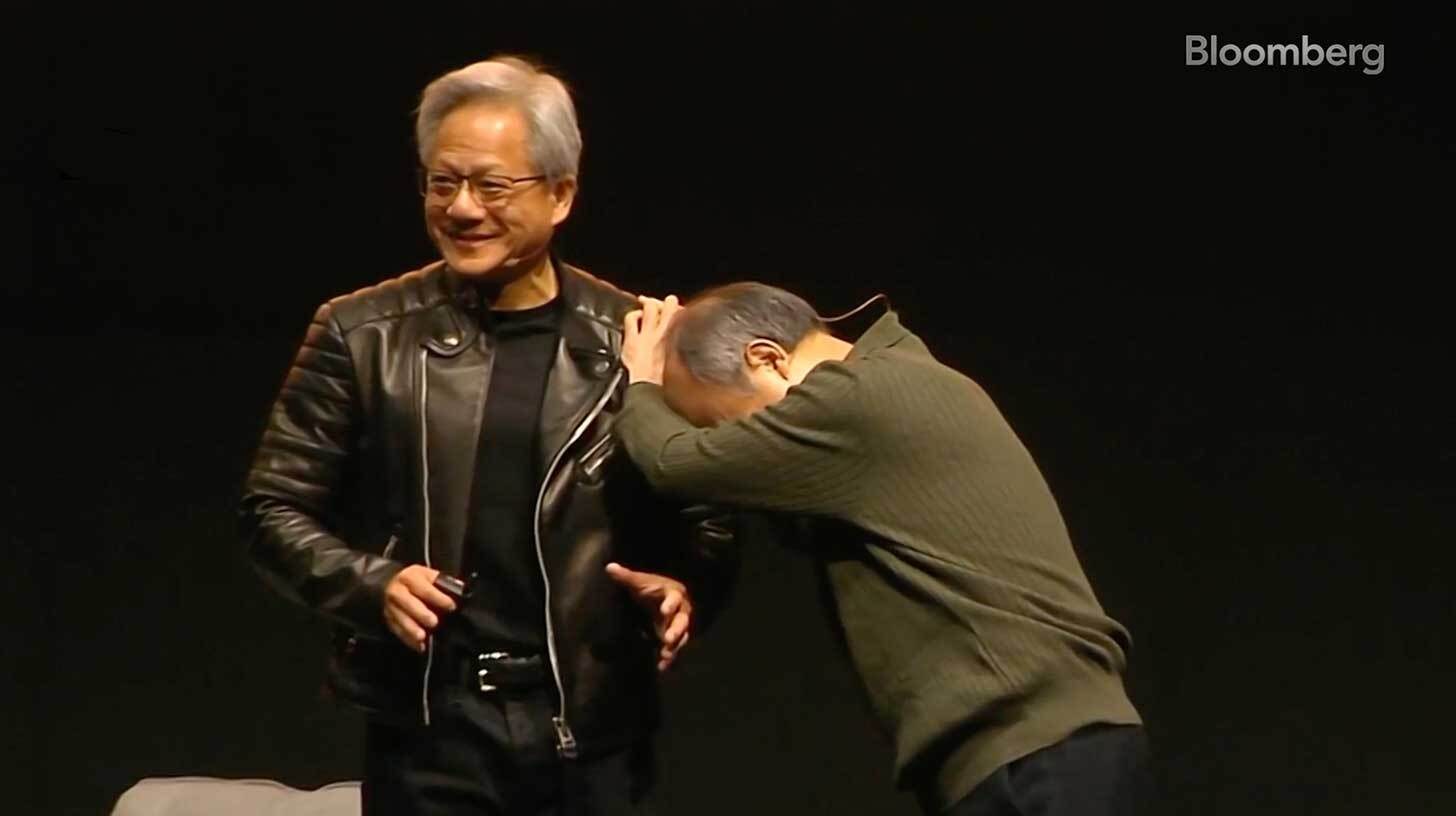 Kaks meest laval, üks neist naeratab ja kannab musta nahkjopet, teine kummardab ja hoiab tema kätt. Taustal on must sein ja tekst 'Bloomberg' on üleval paremas nurgas.