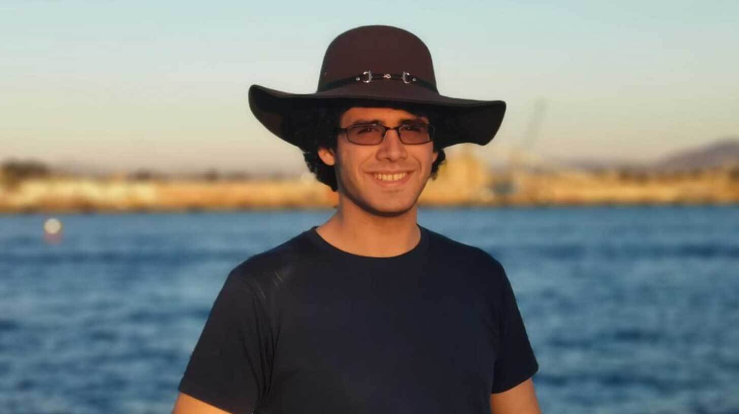 Mees seisab õues vee ees, kandes laia äärega musta kübarat ja tumedaid prille. Tal on seljas tumesinine särk ja ta naeratab. Taustal on näha veekogu ja udune maa-ala.