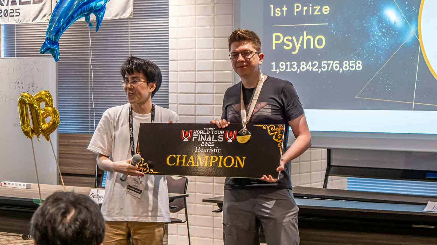 Kahe meheline rühm seisab koos ja hoiab käes suurt musta tahvlit kirjaga 'World Tour Finals 2025 Heuristic Champion'. Üks neist hoiab mikrofoni ja teine kannab medalit. Taustal ekraanil on tekst '1st Prize Psyo 1,913,842,576,858'. Vasakul küljel on näha sinine õhupall ja number '10B' kuldsetes õhupallides.